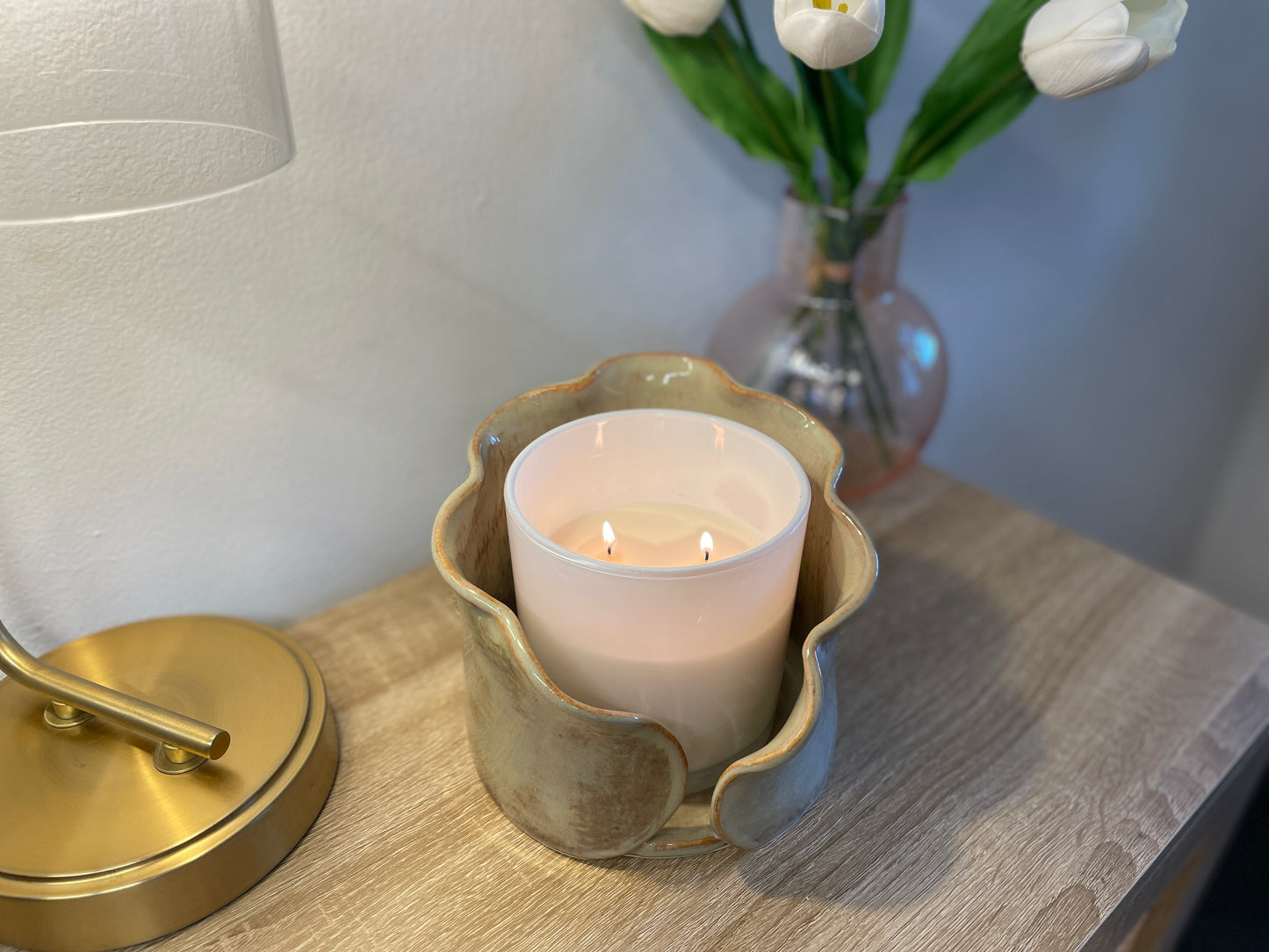 Candle in a decorative ceramic centerpiece holder on a wooden surface with a lamp and flowers in the background