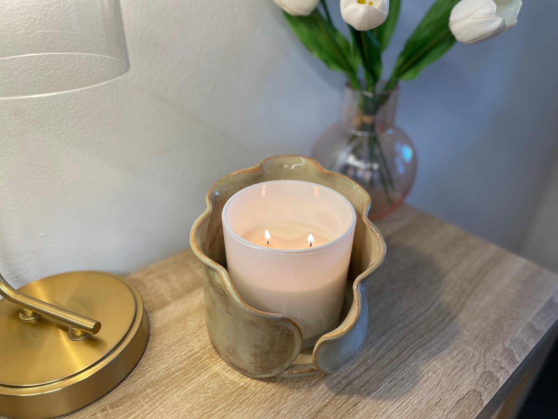 Candle in a decorative ceramic centerpiece holder on a wooden surface with a lamp and flowers in the background