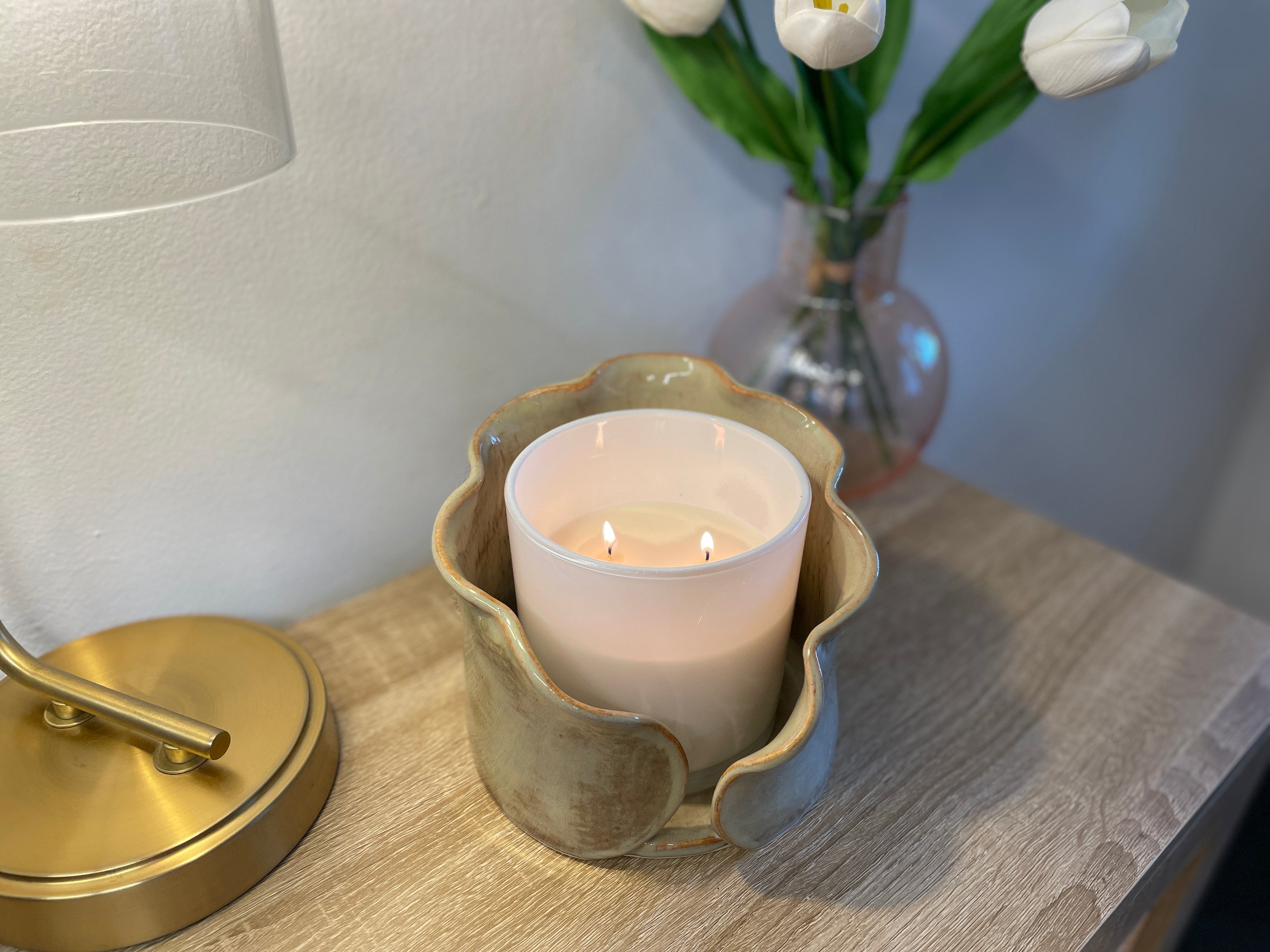 Candle in a decorative ceramic centerpiece holder on a wooden surface with a lamp and flowers in the background