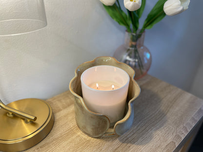 Candle in a decorative ceramic centerpiece holder on a wooden surface with a lamp and flowers in the background